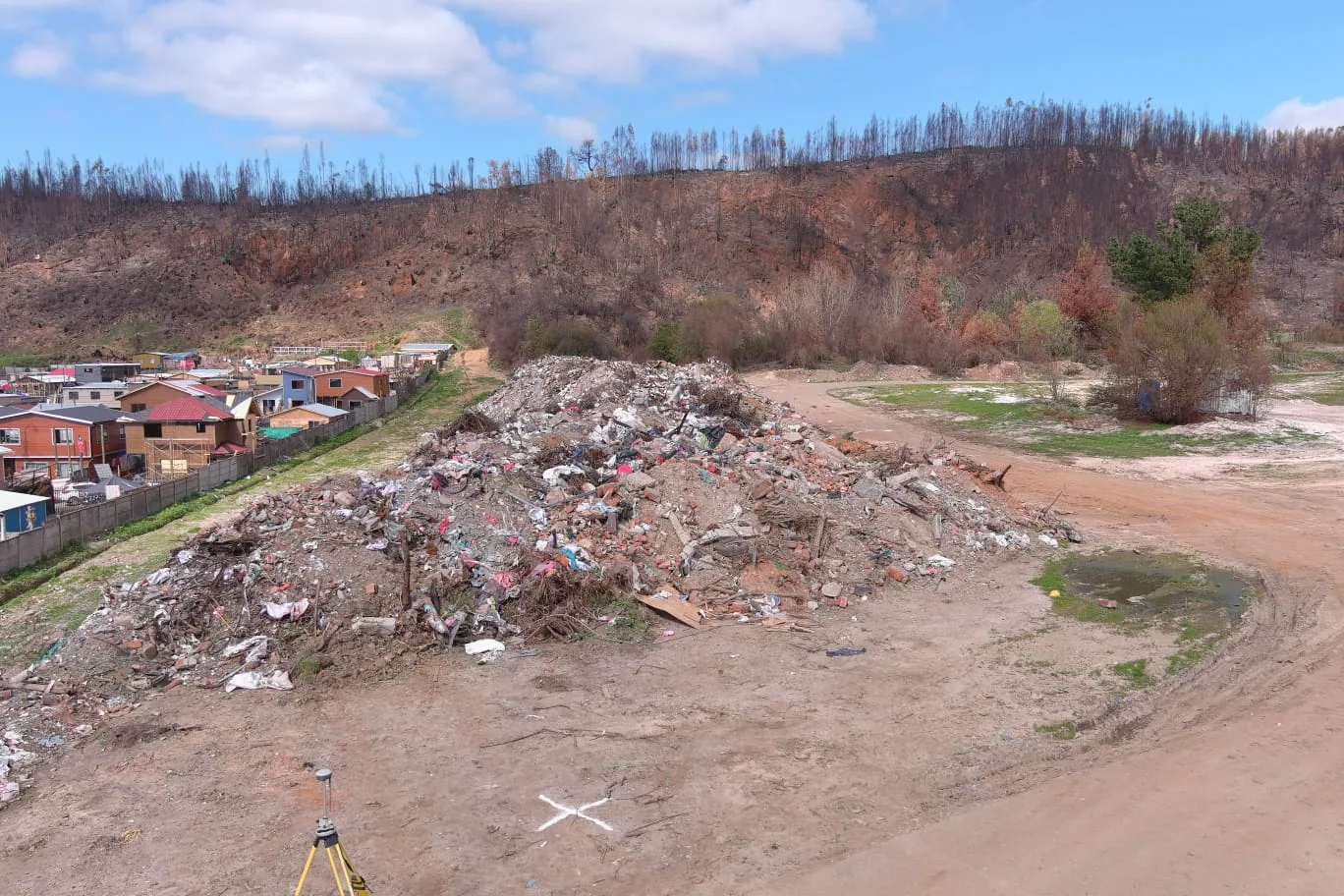 Receptor GPS RTK sobre trípode en terreno, frente a un acopio de escombros y una ladera afectada por incendio
